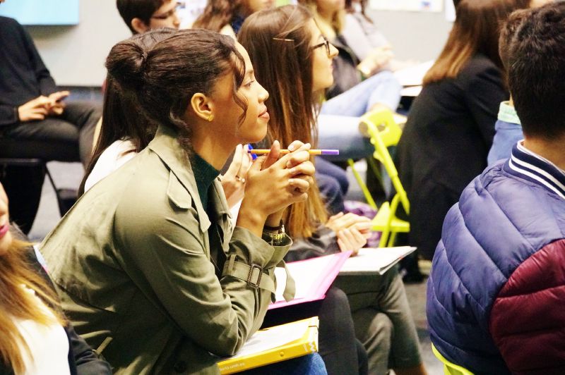 En un aula magna semicircular una persona está hablando y en los pupitres otras personas escuchando