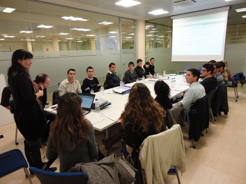 Una chica de pie delante de una mesa repleta de personas sentadas en una oficina con una pantalla al fondo Practicas