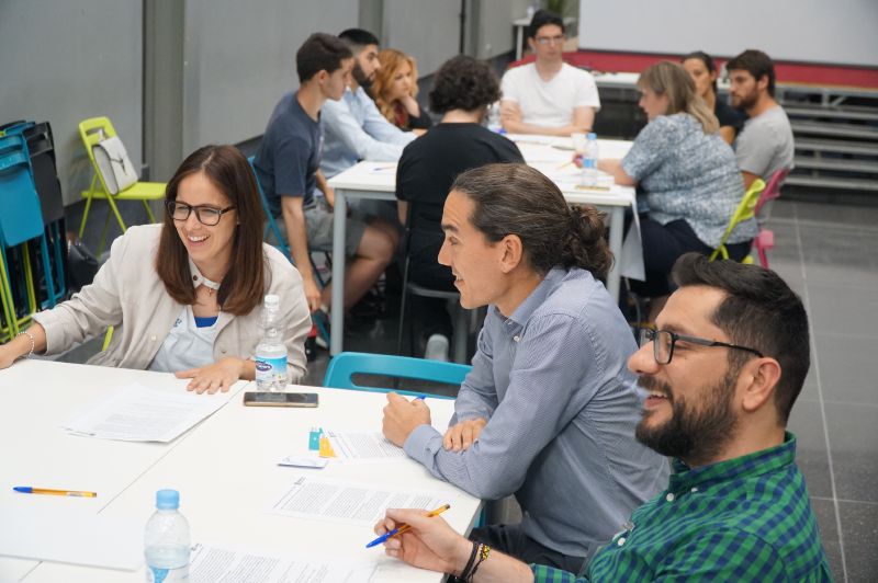 En un aula magna semicircular una persona está hablando y en los pupitres otras personas escuchando