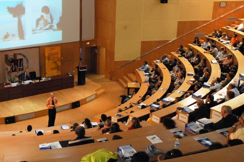 En un aula magna semicircular una persona está hablando y en los pupitres otras personas escuchando